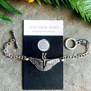Pode incluir: Um bracelete de corrente de prata com um fecho de borboleta detalhado. O bracelete tem um fecho de correr e um anel de prata com um logotipo que diz "Head Turner Studio Joias artesanais".