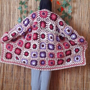 May include: A handmade, open-front crochet cardigan in a granny square pattern. The cardigan features a color palette of pink, purple, and cream squares. The text "Handmadebybircool" is visible on the cardigan. The background is a bamboo wall.