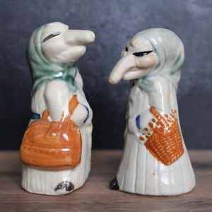 May include: A pair of vintage ceramic figurines depicting elderly figures with long noses and hooded head coverings. One figure holds an orange bag, the other a basket. The figures are painted in cream, green, and orange.