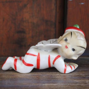 May include: A ceramic figurine of a crawling elf with white skin, wearing a white suit featuring horizontal red stripes. The elf has small white wings on its back and a red hat with a green pom-pom on top. The face has painted features.