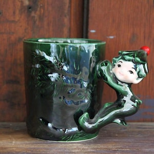May include: A dark green ceramic mug with a sculpted elf handle. The mug features a textured design of evergreen trees. The elf handle has a smiling face and a red berry on its hat. A unique holiday-themed coffee mug.