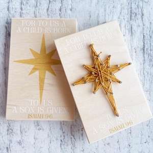 May include: Two rectangular light wood plaques featuring the text FOR TO US A CHILD IS BORN TO US A SON IS GIVEN ISAIAH 9:6. One plaque displays a flat gold star graphic, while the other features a three-dimensional star made of gold string and nails.