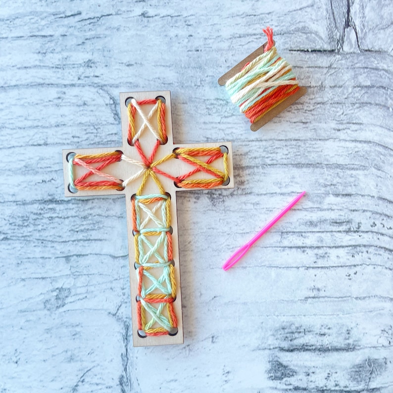 May include: A wooden cross with a colorful string design. The cross is on a white wooden surface with a spool of string and a pink needle.