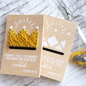 May include: Two wooden plaques with the text "PERHAPS THIS IS THE MOMENT FOR WHICH YOU HAVE BEEN CREATED. ESTHER 4:14". One plaque features a yellow string art crown. The other has a white geometric design. A clear bag contains yellow string and small nails.
