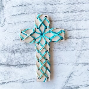 May include: A wooden cross with a blue and green yarn design. The yarn is woven in a criss-cross pattern.