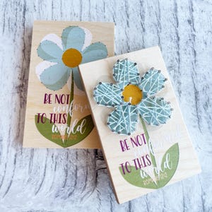 Op de afbeelding: Twee rechthoekige houten platen met een lichtblauw bloemmotief en een geel hart. De tekst luidt BE NOT conformed TO THIS world ROMANS 12:2. Eén plaat heeft gedrukte afbeeldingen, terwijl de andere is voorzien van lichtblauwe draadkunst.