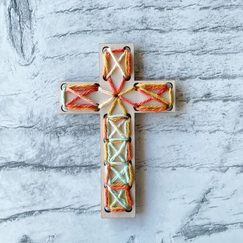 May include: A wooden cross with a colorful thread design. The cross is decorated with orange, yellow, white, and light blue thread.