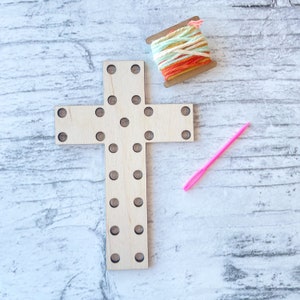 May include: A wooden cross with holes for stringing yarn or thread. The cross is light brown and has a pink needle and a spool of colorful yarn next to it.
