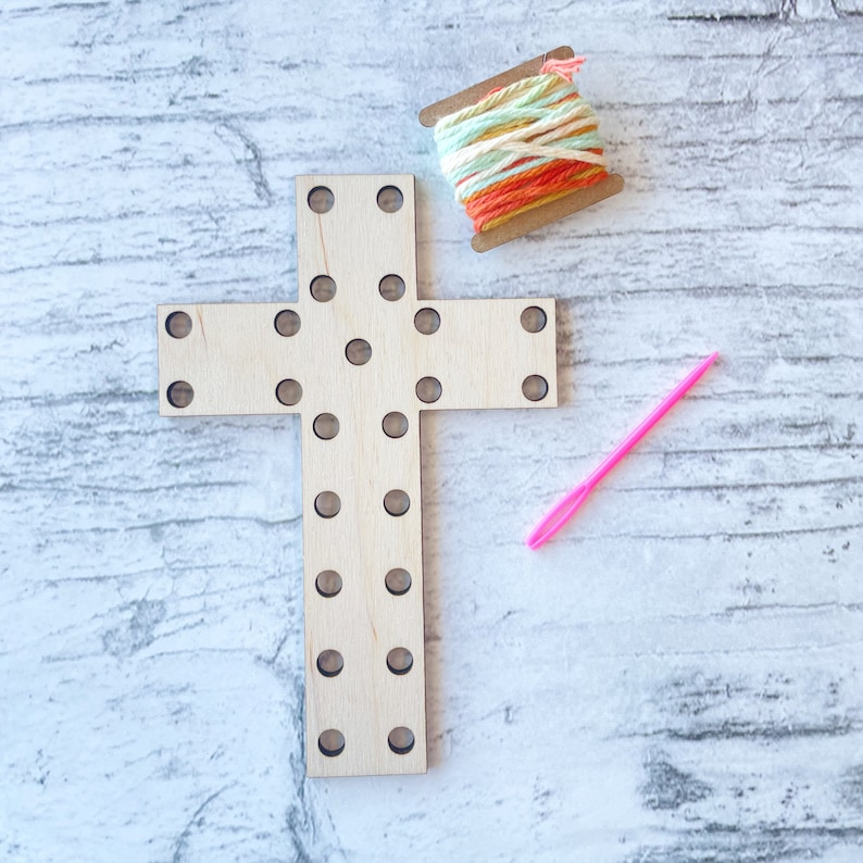 May include: A wooden cross with holes for stringing yarn or thread. The cross is light brown and has a pink needle and a spool of colorful yarn next to it.