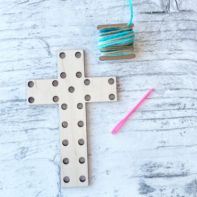 May include: A wooden cross with holes for stringing yarn. The cross is light brown and has a pink needle and a spool of blue and green yarn next to it.
