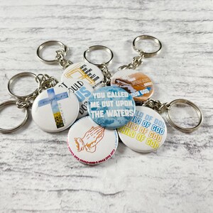 Könnte beinhalten: Sechs bunte Schlüsselanhänger mit verschiedenen religiösen Botschaften. Die Schlüsselanhänger zeigen ein Kreuz, betende Hände und den Spruch "Du hast mich auf die Wasser gerufen".
