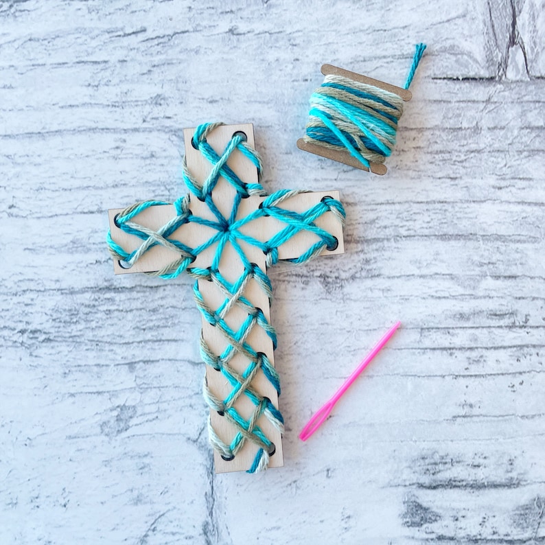 May include: A wooden cross with blue and green yarn woven into a pattern. A spool of yarn and a pink needle are also included.