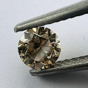 May include: A round, light brown diamond gemstone held by tweezers. The diamond has a faceted cut and is clear with a slight yellow hue.