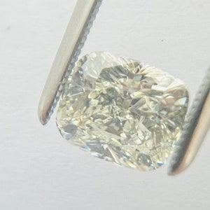 May include: A cushion-cut diamond held by silver tweezers. The diamond has a light yellow hue and a faceted surface that reflects light. The shape is rectangular with rounded corners, and the facets create a sparkling effect.