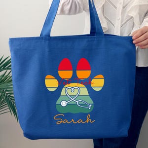 May include: A blue tote bag with a paw print design featuring the words "Vet Tech", a stethoscope, and the name "Sarah". The paw print has orange, yellow, and green accents. The bag has two blue handles.