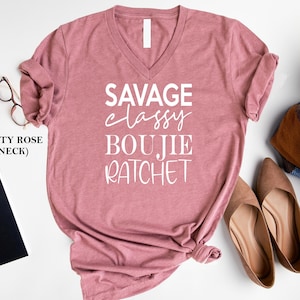 May include: Dusty rose V-neck t-shirt with the text "Savage Classy Boujie Ratchet" printed in white.