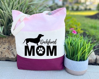May include: A tri-color tote bag with a pink, white, and burgundy design. The bag features a black silhouette of a dachshund and the text "Dachshund MOM" with a paw print.