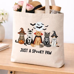 May include: A beige canvas tote bag featuring a graphic of five cats in Halloween costumes, including a witch, mummy, vampire, skeleton, and wizard, with a full moon, bats, and the text JUST A SP**KY PAW printed in black ink on the front.