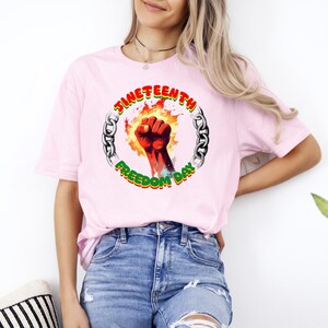 May include: A light pink t-shirt featuring a graphic celebrating Juneteenth. The design includes a red fist, flames, and the words "Juneteenth Freedom Day" in a circular chain design. The shirt is made of a soft material.