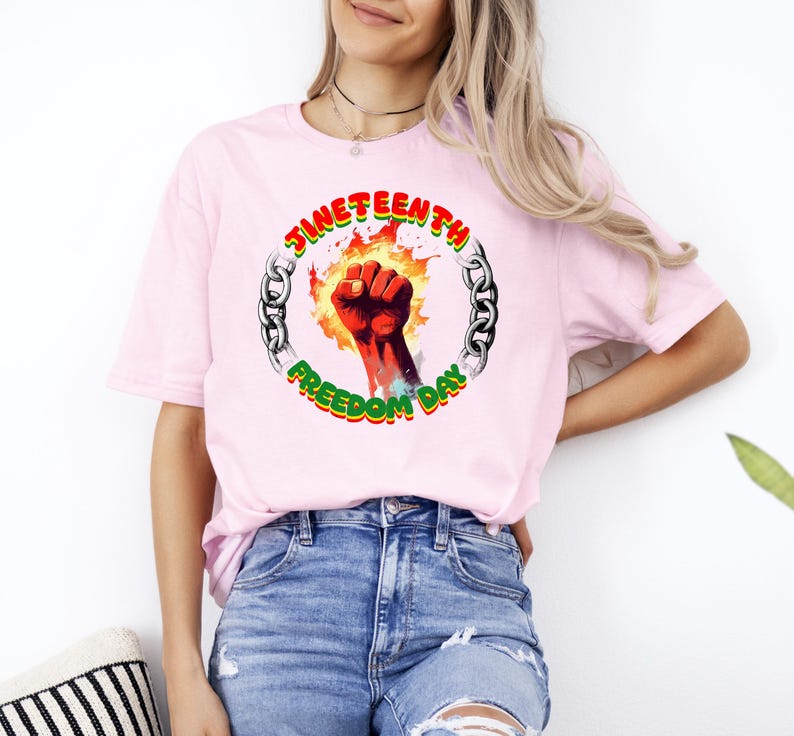 May include: A light pink t-shirt featuring a graphic celebrating Juneteenth. The design includes a red fist, flames, and the words "Juneteenth Freedom Day" in a circular chain design. The shirt is made of a soft material.
