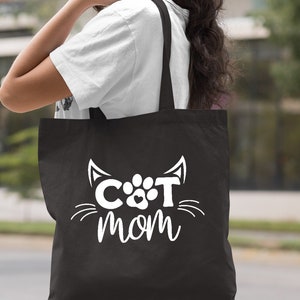 Pode incluir: Uma bolsa tote preta com texto branco que diz "CAT MOM" com uma pegada de pata e orelhas de gato.
