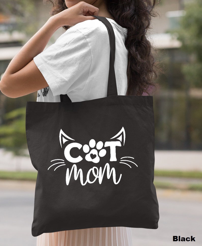 Pode incluir: Uma bolsa tote preta com texto branco que diz "CAT MOM" com uma pegada de pata e orelhas de gato.