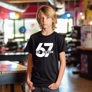 May include: A black t-shirt with the number "67" in white, with the words "SIX" and "SEVEN" below. The shirt is being worn by a person with blond hair.
