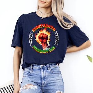 May include: Navy blue t-shirt featuring a circular graphic with the text "Juneteenth Freedom Day." The design includes a raised fist in red, surrounded by flames and a chain border. The text is in green and yellow.