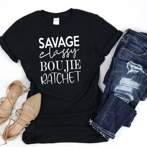 May include: Black t-shirt with white text that reads "Savage Classy Boujie Ratchet". The shirt is paired with blue jeans and brown sandals.