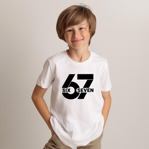 May include: A white short-sleeved t-shirt with the number "67" in a bold black font. The words "SIX" and "SEVEN" are printed below the number. The shirt is worn by a person.