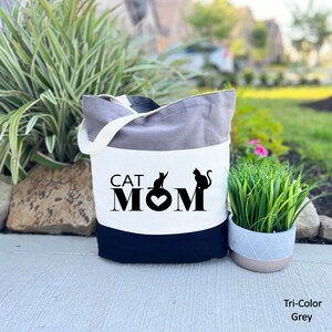 Puede incluir: Una bolsa de mano tricolor con un panel blanco que presenta el texto "CAT MOM" en negro con dos siluetas de gatos negros y un coraz&oacute;n. La bolsa es gris, blanca y negra.
