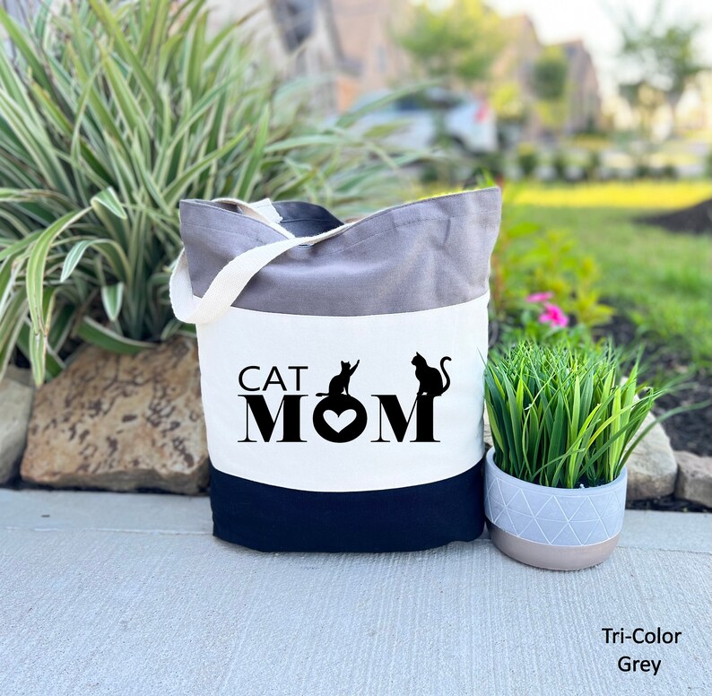 Puede incluir: Una bolsa de mano tricolor con un panel blanco que presenta el texto "CAT MOM" en negro con dos siluetas de gatos negros y un coraz&oacute;n. La bolsa es gris, blanca y negra.