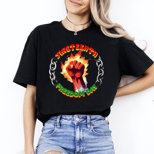 May include: Black t-shirt featuring a circular graphic with the text "Juneteenth Freedom Day." The graphic includes a red fist, flames, and a chain. The text is in green and yellow. The shirt is made of a soft material.