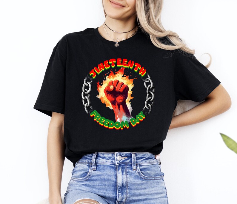 May include: Black t-shirt featuring a circular graphic with the text "Juneteenth Freedom Day." The graphic includes a red fist, flames, and a chain. The text is in green and yellow. The shirt is made of a soft material.
