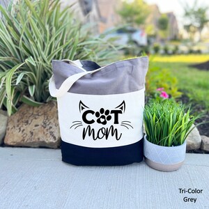 Pode incluir: Uma bolsa tote tricolor com um painel branco que apresenta um rosto de gato preto com uma pegada de pata como nariz e o texto "CAT MOM". A bolsa &eacute; cinza, branca e preta.