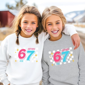 May include: Two young girls wearing matching sweatshirts. The sweatshirts are white and gray, each with a graphic that says "SEVEN SIX 67" in pink and blue, with a checkered background and star accents.