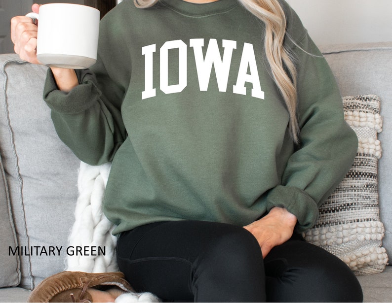 May include: A woman wearing a military green sweatshirt with the word "IOWA" printed in white on the front. She is sitting on a couch and holding a white mug.