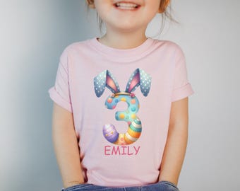 Könnte beinhalten: Rosa T-Shirt mit einer bunten Zahl 3, die mit Ostereiern und Hasenohren verziert ist. Der Name "EMILY" ist unter der Zahl gedruckt.
