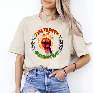 May include: Beige t-shirt featuring a graphic design for Juneteenth. The design includes a red fist surrounded by flames, a chain, and the words "Juneteenth Freedom Day" in green and red.