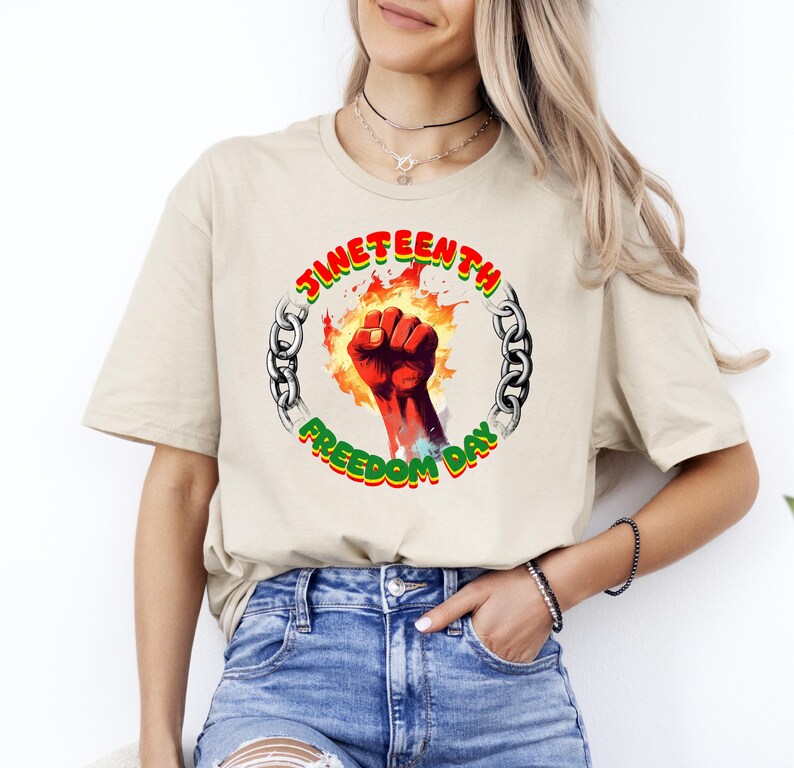 May include: Beige t-shirt featuring a graphic design for Juneteenth. The design includes a red fist surrounded by flames, a chain, and the words "Juneteenth Freedom Day" in green and red.