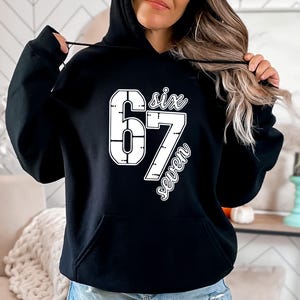 May include: Black hooded sweatshirt with a white graphic that reads "six 67 seven". The numbers are in a bold, blocky font, and the words are in a script font. The sweatshirt has a front pocket and a drawstring hood.