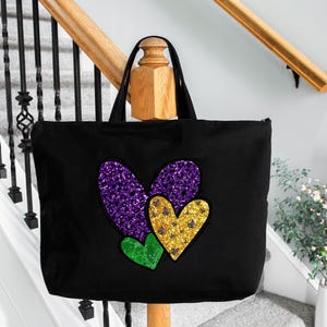 Puede incluir: Una bolsa de lona negra con tres corazones brillantes. Los corazones son morados, dorados y verdes. La bolsa est&aacute; sobre una barandilla de madera.
