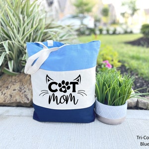 Pode incluir: Uma bolsa tote tricolor com listras azuis, brancas e azul marinho. A bolsa tem um painel branco com o texto "CAT MOM" em letras pretas e uma pegada no meio do "O".