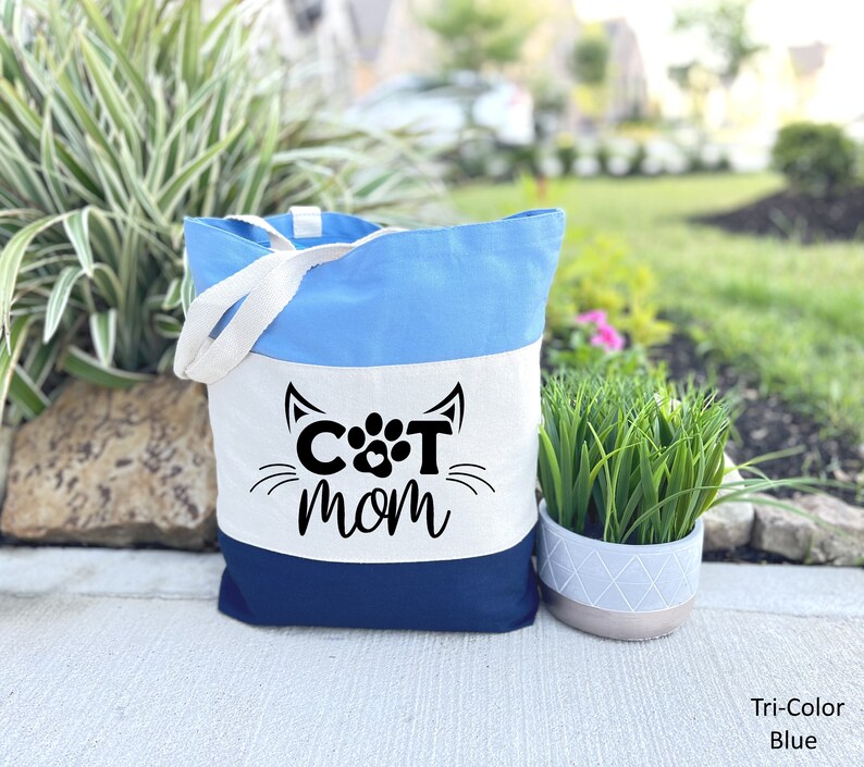 Pode incluir: Uma bolsa tote tricolor com listras azuis, brancas e azul marinho. A bolsa tem um painel branco com o texto "CAT MOM" em letras pretas e uma pegada no meio do "O".