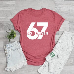 May include: A red t-shirt with the number "67" in white, with the words "SIX SEVEN" below the number. The shirt is paired with denim shorts, a white sweater, and a coffee cup.