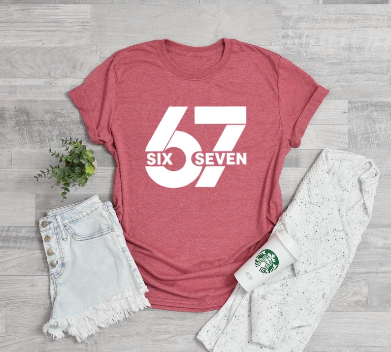 May include: A red t-shirt with the number "67" in white, with the words "SIX SEVEN" below the number. The shirt is paired with denim shorts, a white sweater, and a coffee cup.