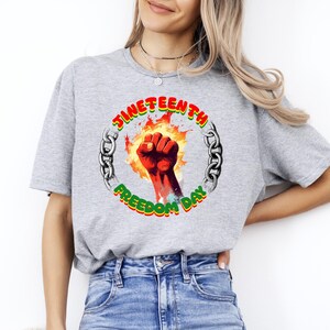 May include: A gray t-shirt featuring a graphic for Juneteenth Freedom Day. The design includes a red fist emerging from flames, surrounded by a chain and the words "Juneteenth Freedom Day" in red, green, and yellow.