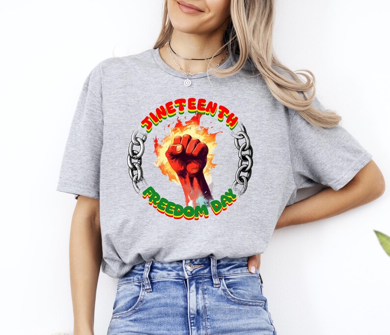 May include: A gray t-shirt featuring a graphic for Juneteenth Freedom Day. The design includes a red fist emerging from flames, surrounded by a chain and the words "Juneteenth Freedom Day" in red, green, and yellow.