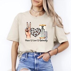May include: Beige t-shirt featuring a peace sign hand gesture, a heart with a leopard print design, and an eye chart. The text "Peace Love Optometry" is printed below the graphics. The shirt is made of a soft material.