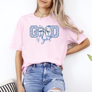 K&ouml;nnte beinhalten: Ein pinkfarbenes T-Shirt mit einem blauen und wei&szlig;en Schleifen-Motiv und dem Text "GOD is good".